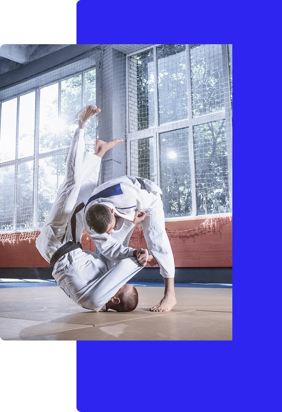 Two martial artists wearing white gis train in a well-lit gym. One performs a judo throw, flipping the other over his shoulder.